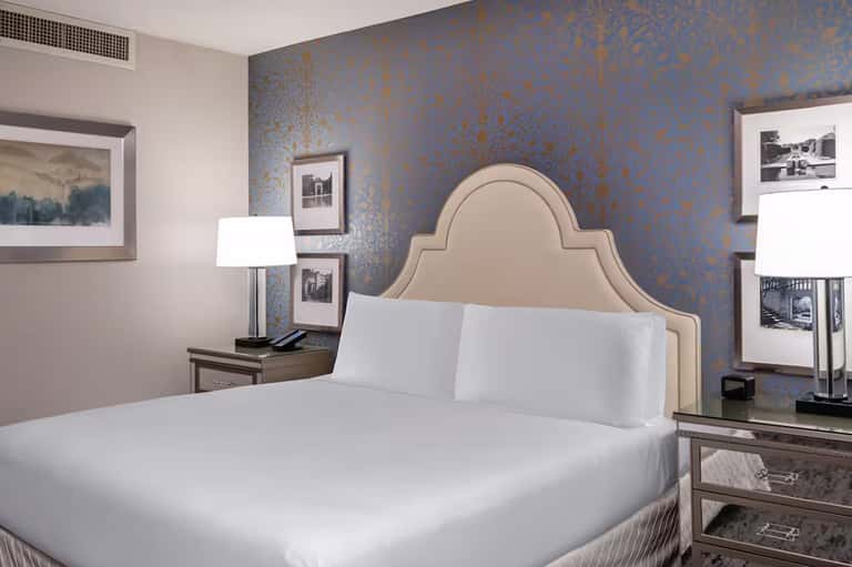 Elegant hotel bedroom with beige upholstered headboard, white bedding, blue damask wallpaper, and modern furnishings