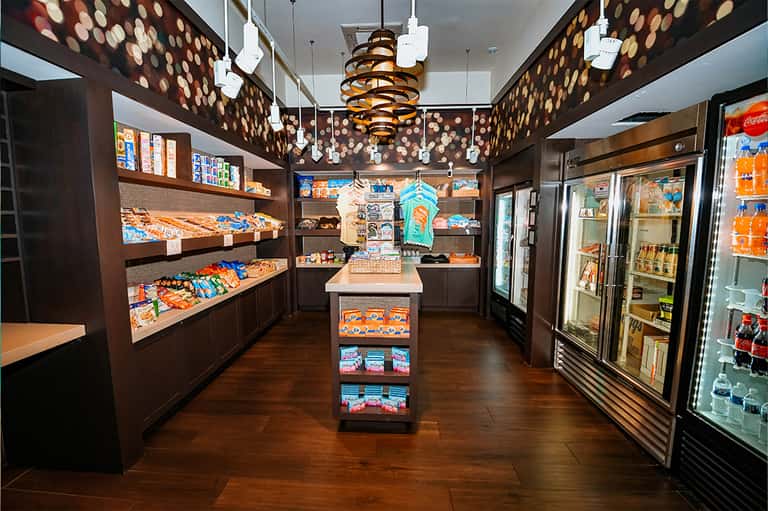 Hotel convenience store interior with dark wood shelving, colorful packaged snacks, refrigerated beverage cases, and pendant lighting