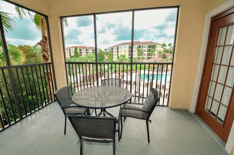 Balcony overlooking resort pool and grounds with round patio table and four chairs in afternoon sunlight.
