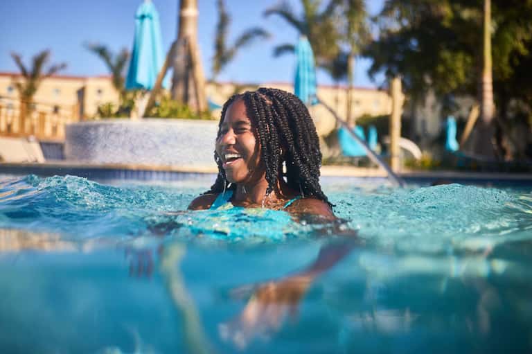 Woman swimming in bright blue resort pool with palm trees and yellow umbrellas in background