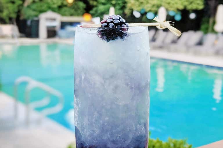 Blackberry cocktail with ice garnished with a gold bow tie, poolside with turquoise water in background