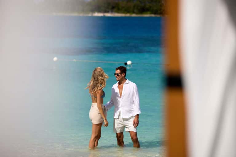 Couple wading in crystal clear turquoise tropical ocean water, framed through white wooden posts