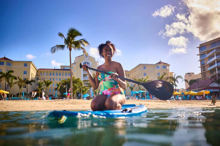 Woman on paddleboard in shallow water near beach with colorful buildings and palm trees in background