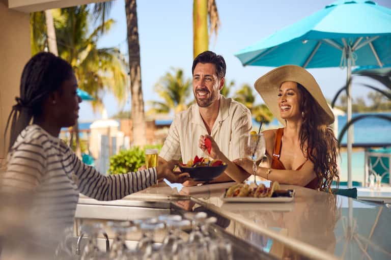 Three people enjoying drinks and fruit at a beachfront bar with palm trees and turquoise umbrellas in background