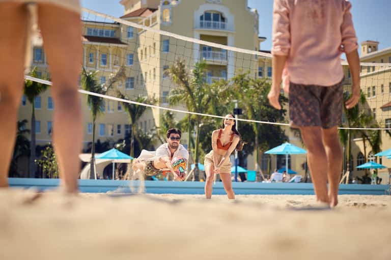 Guests playing beach volleyball on sand with colorful resort buildings and blue umbrellas in background