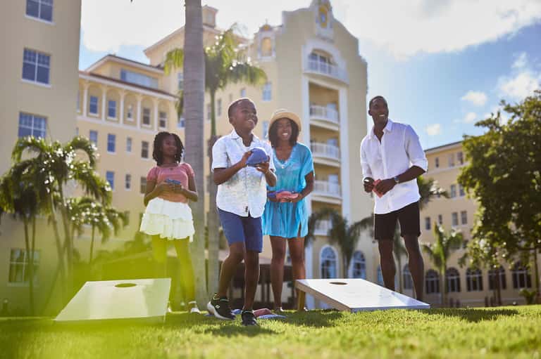 Family playing cornhole outdoors in front of colorful colonial-style resort buildings