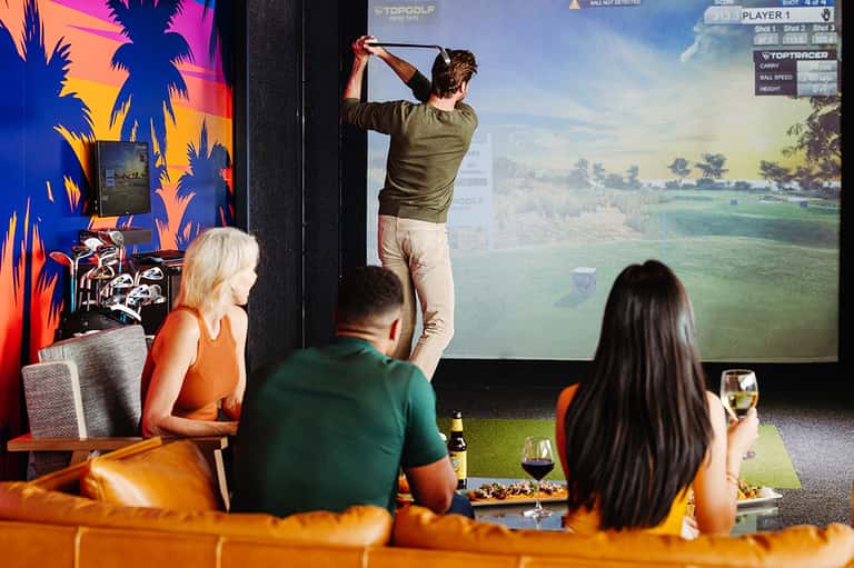 Indoor golf simulator with player swinging club, spectators watching on orange couch with drinks