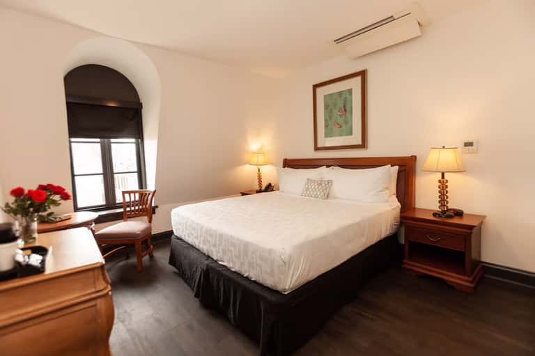 Elegant hotel bedroom with queen bed, wooden furniture, arched window with city view, and warm lighting