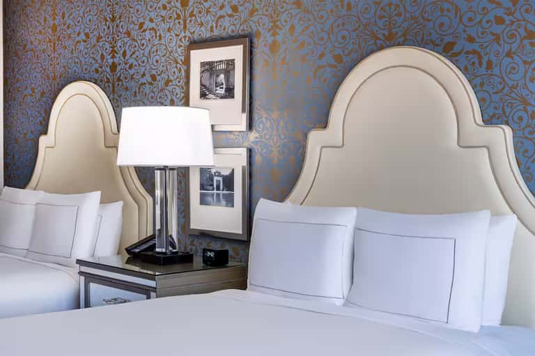 Luxury hotel bedroom with two queen beds, decorative blue and gold damask wallpaper, cream headboards, and framed artwork.