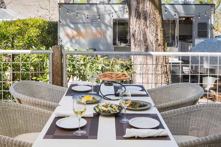Outdoor dining area with wicker chairs, set table with wine glasses, salad dishes, and farmhouse restaurant in background