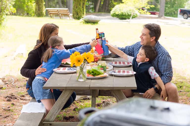 Family of four toasting with drinks at outdoor picnic table with yellow flowers and food