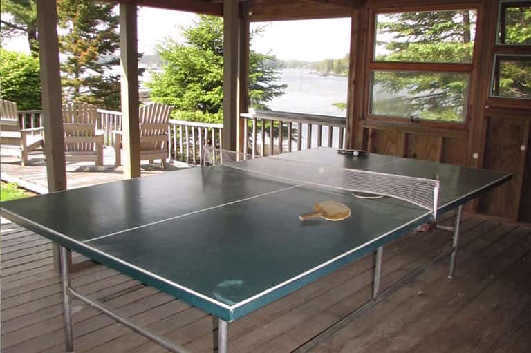 Covered deck overlooking a lake with ping pong table, wooden chairs, and scenic water views surrounded by trees