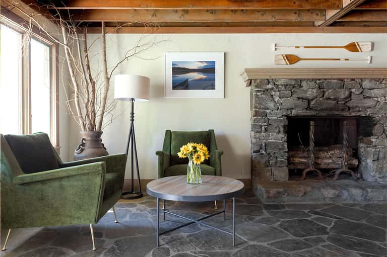 Rustic lounge with green chairs, fireplace, and stone hearth facing large windows with natural branches in vase