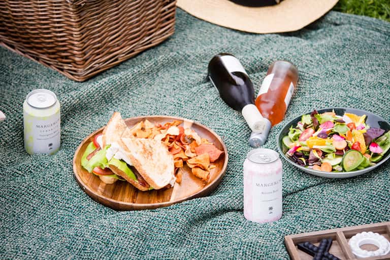 Outdoor picnic setup with sandwich, colorful salad, wine bottles, and Margeré branded beverages on teal textured surface