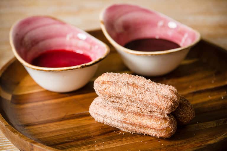 Cinnamon sugar-coated pastries with pink bowls of red dipping sauce on wooden board
