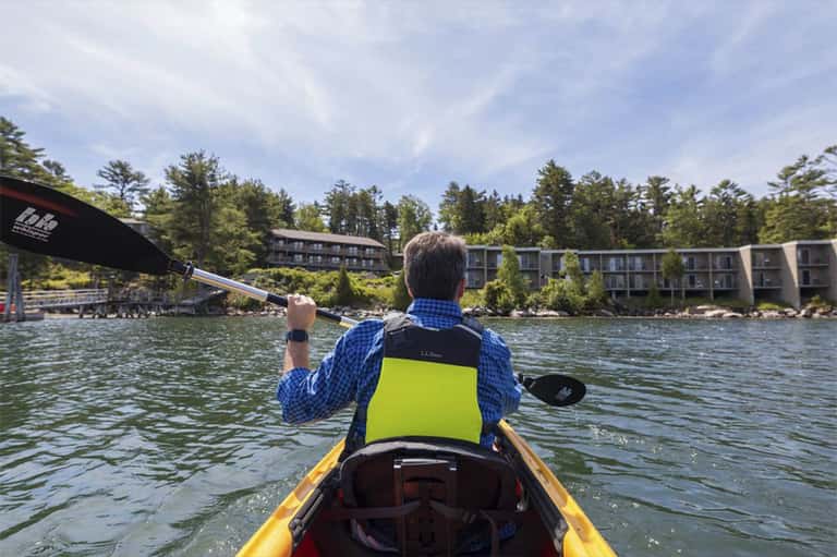 Man kayaking in yellow kayak on calm lake with residential buildings and forest in background