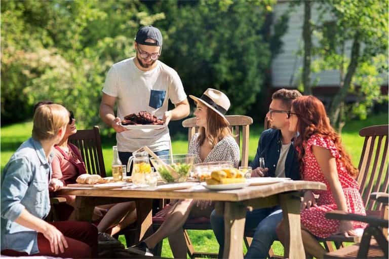 Family enjoying outdoor meal at wooden table with food and drinks in sunny garden setting
