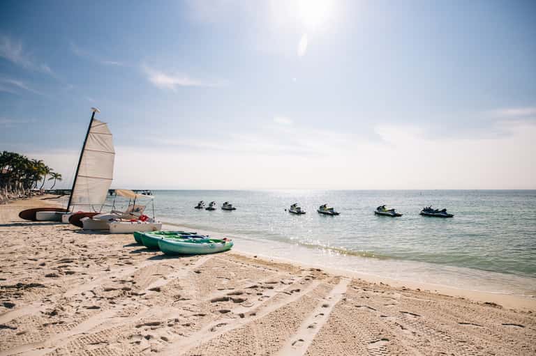 Sandy beach with sailboat and green kayak, jet skis visible in turquoise ocean water