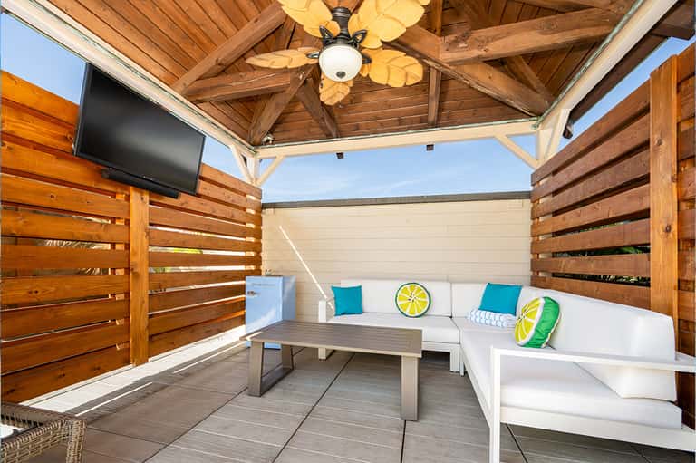 Covered wooden deck lounge with white seating, turquoise pillows, ceiling fan, and tropical outdoor space