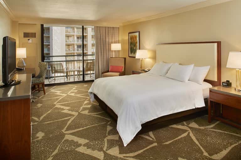 Spacious hotel bedroom with king bed, cream bedding, wood furnishings, and balcony overlooking city buildings