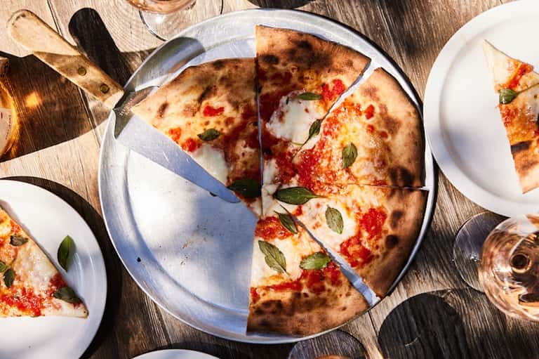 Freshly baked Margherita pizza on white plate with basil, surrounded by additional slices on wooden table