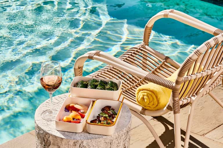 Poolside lounge chair with rose wine, gourmet food containers, and yellow towel by sparkling water
