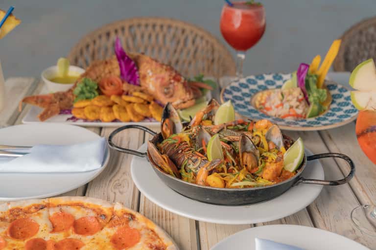Beachfront dining table with seafood paella, grilled fish, and cocktails with colorful garnishes