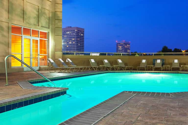 Turquoise rooftop pool at night with lounge chairs and city buildings in background