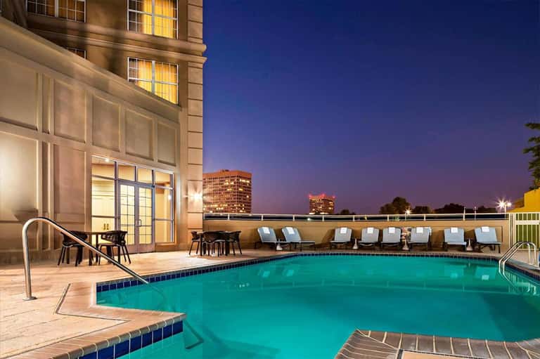 Illuminated resort pool at dusk with lounge chairs, stone waterfall feature, and high-rise building backdrop