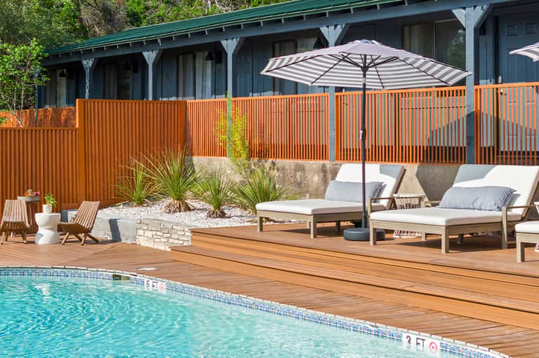 Resort pool deck with wooden loungers, striped umbrella, modern house with orange railing, and lush landscaping