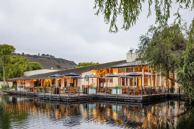 Modern waterfront restaurant with wooden deck, large windows, and blue umbrellas overlooking a river