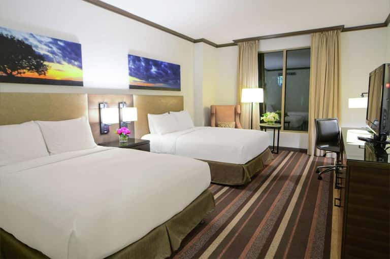 Hotel room with two queen beds, tan headboards, artwork, desk area, and balcony with curtains