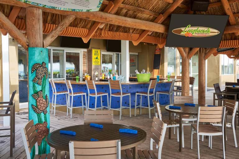 Tropical beachfront bar with thatched roof, blue cushioned seating, and wooden tables under covered patio