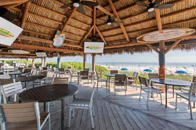 Beachfront tiki bar with wooden thatched roof, dining tables, ocean view, and colorful umbrellas on sand