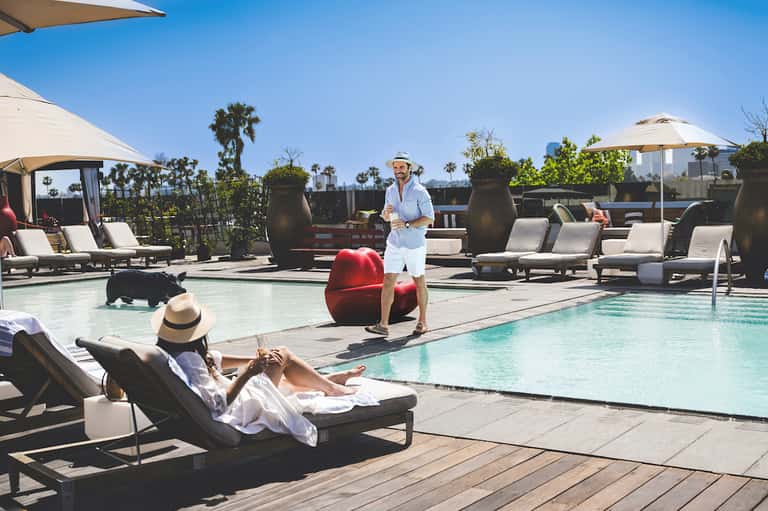 Resort rooftop pool with lounge chairs, umbrellas, palm trees, and city skyline views on a sunny day