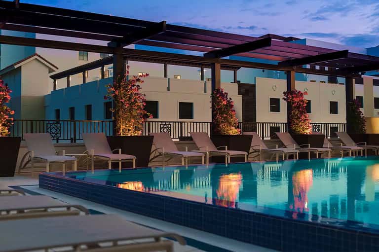 Contemporary pool deck at dusk with lounge chairs, pergola, and illuminated building facade