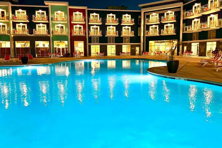 Brightly lit resort pool at night reflecting colorful building facades with balconies and lounge chairs