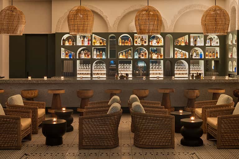 Upscale bar lounge with woven pendant lights, wicker seating, dark shelving with liquor bottles, and ambient candlelit tables