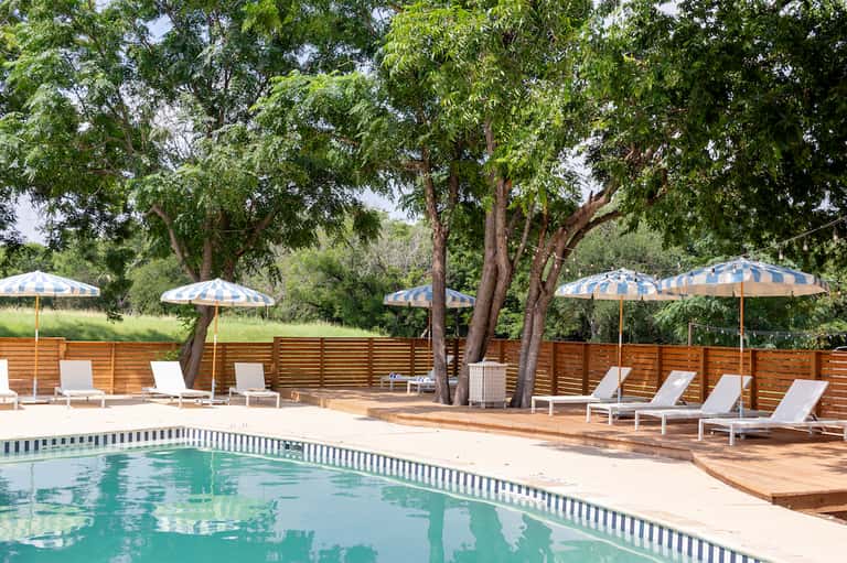 Resort pool with lounge chairs, blue and white umbrellas, and tall shade trees with wooden fence backdrop
