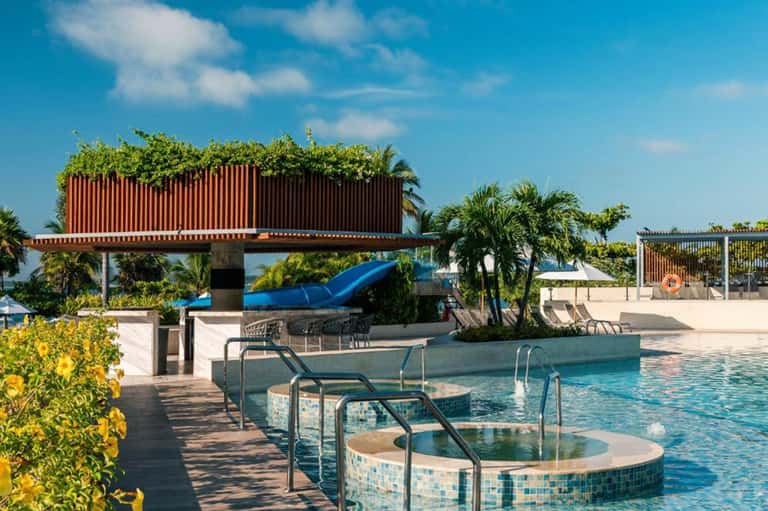 Resort pool area with water slide, hot tubs, and tropical landscaping under blue sky