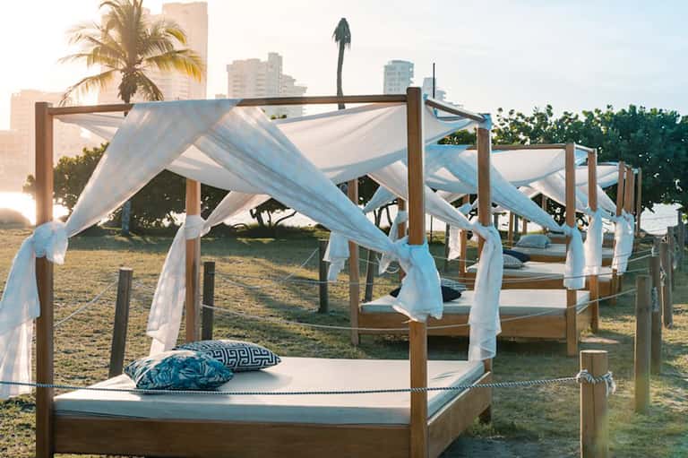 Beach cabana bed with white sheer curtains and blue pillows at sunset with city skyline backdrop