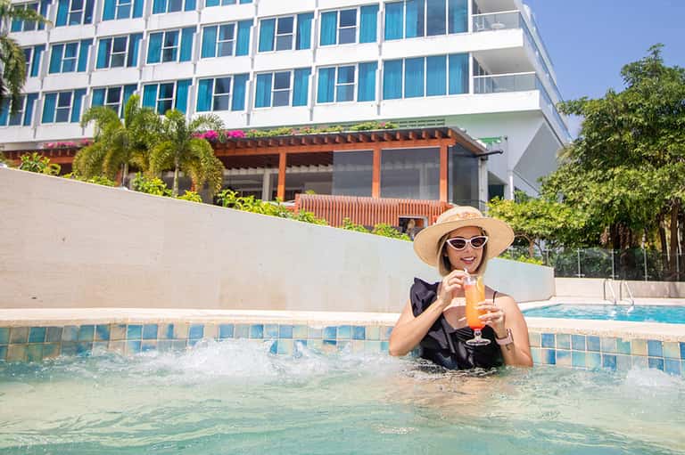 Woman in a pool at a modern resort holding a tropical drink, wearing a hat and sunglasses