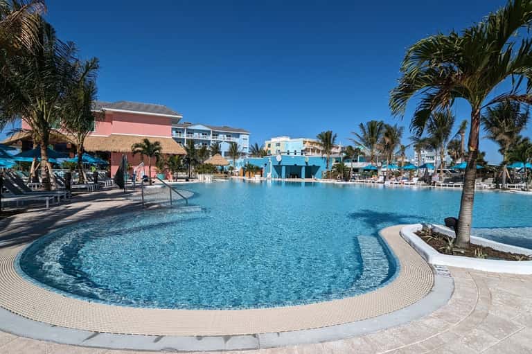 Resort pool area with turquoise water, palm trees, and pastel-colored buildings