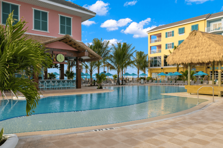 Resort pool area with colorful buildings, tiki bar, palm trees, and beach access visible