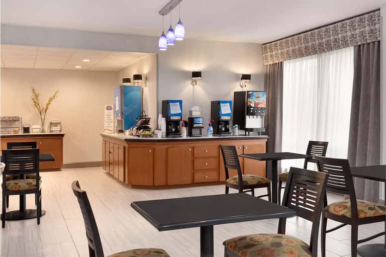 Modern hotel lobby with wooden reception desk, beverage station, and dining area with pendant lighting