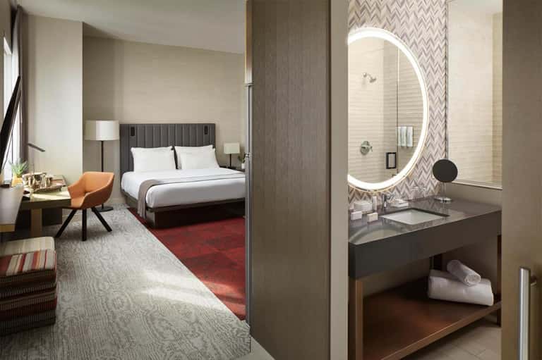 Modern hotel room with king bed, ensuite bathroom with illuminated round mirror, and contemporary furnishings