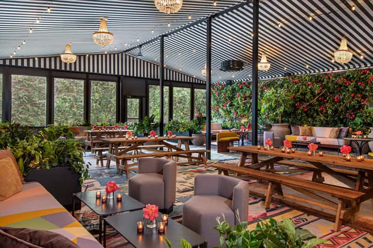 Elegant rooftop lounge with black striped pergola, wooden furniture, flowering plants, and crystal chandeliers