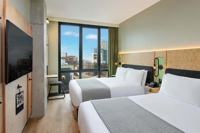 Modern hotel room with two queen beds, floor-to-ceiling windows overlooking city skyline, and contemporary furnishings