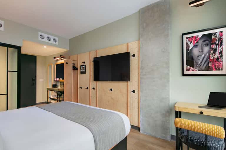 Modern hotel bedroom with plywood storage walls, flat-screen TV, desk with laptop, and contemporary furnishings