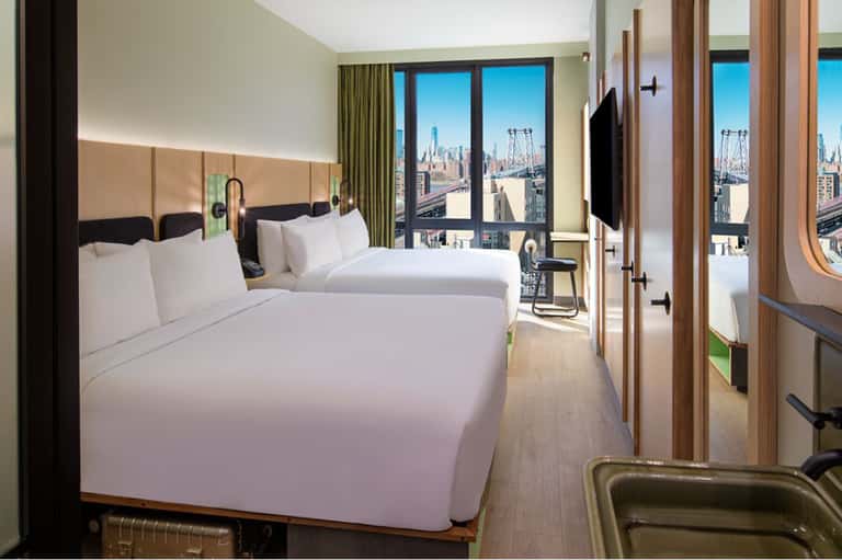 Luxury hotel bedroom with two queen beds, green curtains, and expansive windows overlooking city skyline